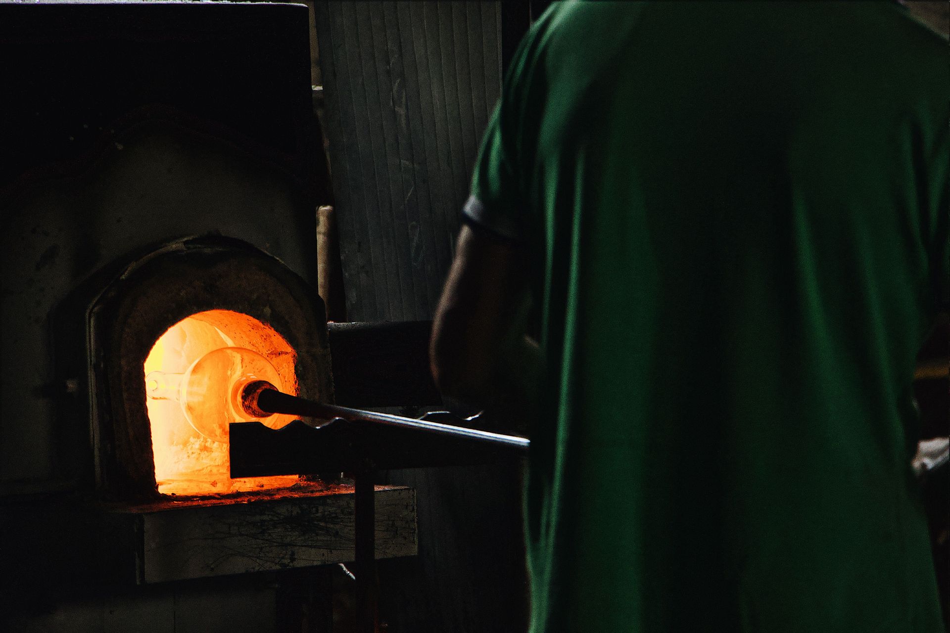 Back view of crop unrecognizable professional worker standing in front of furnace while blowing glass in workshop